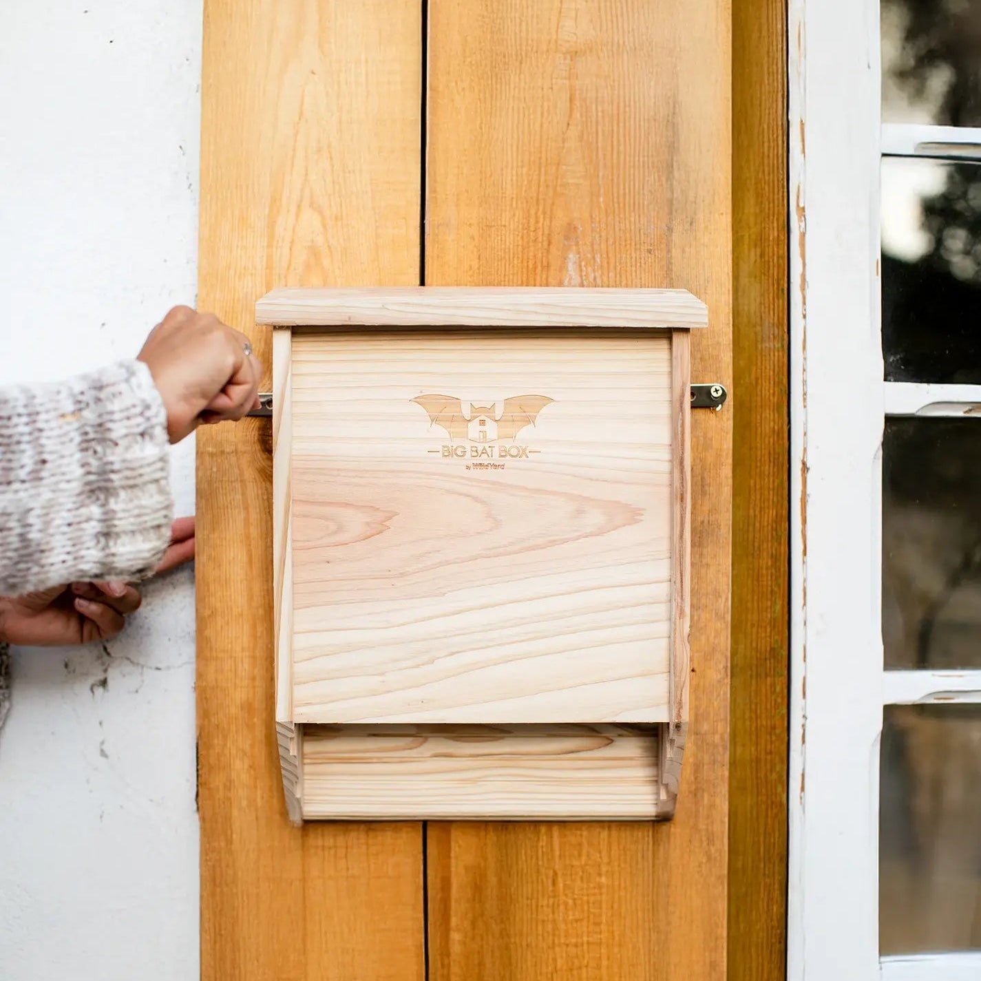 Cedar two-chamber bat house attached to a wooden wall with a blurred window in the background.